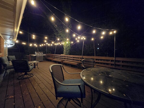 Terraza al aire libre con luces colgantes, bancos de madera y una parrilla.