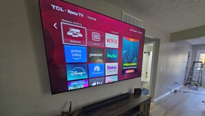 A TCL Roku TV mounted on a wall with a wooden entertainment center below.
