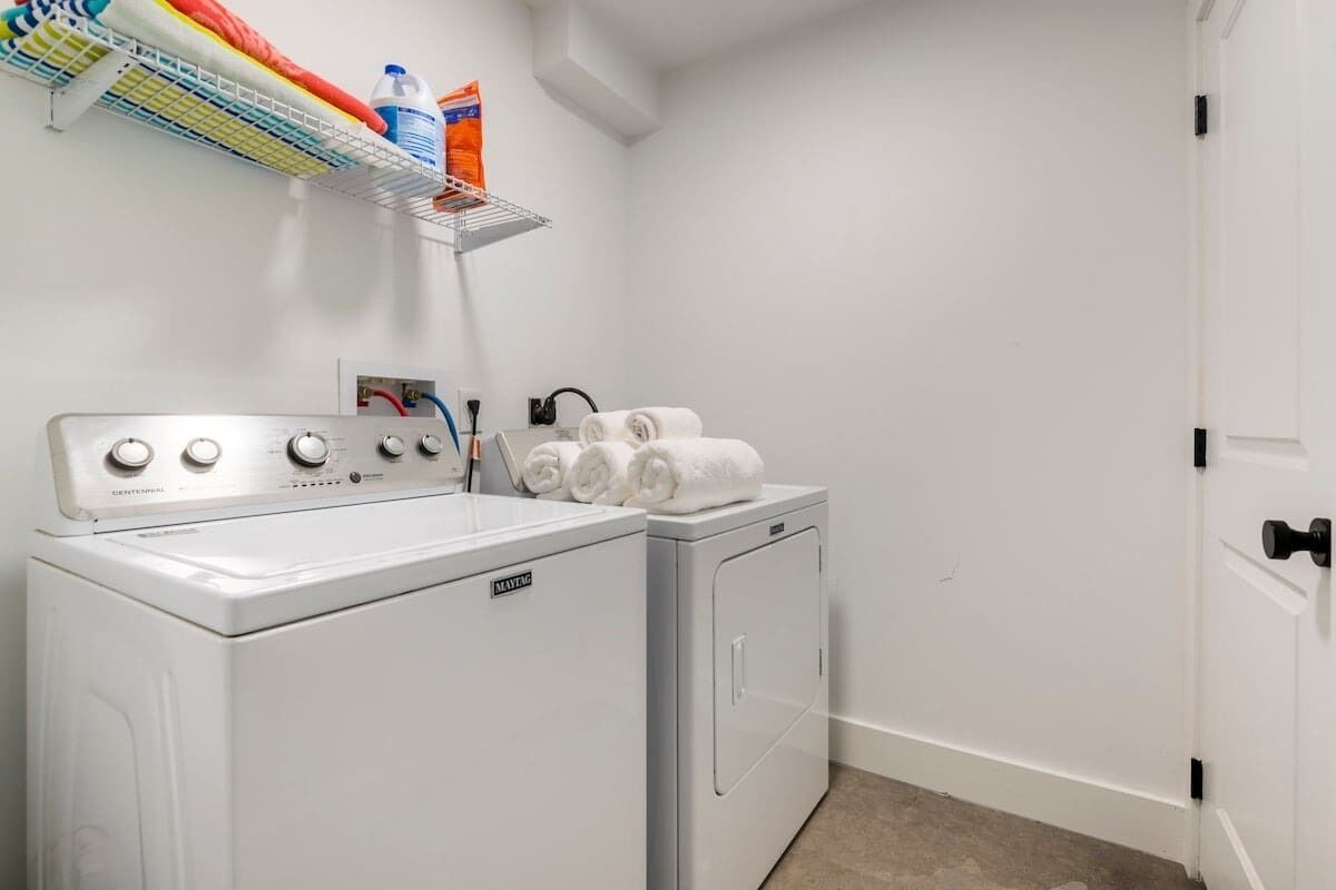 A laundry area with a front-loading washer and dryer, a shelf with laundry supplies, and a door to the right.