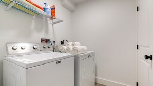A laundry area with a front-loading washer and dryer, a shelf with laundry supplies, and a door to the right. - Gallery House: Designer Home-Hot Tub-Campfire-EV (Ithaca)