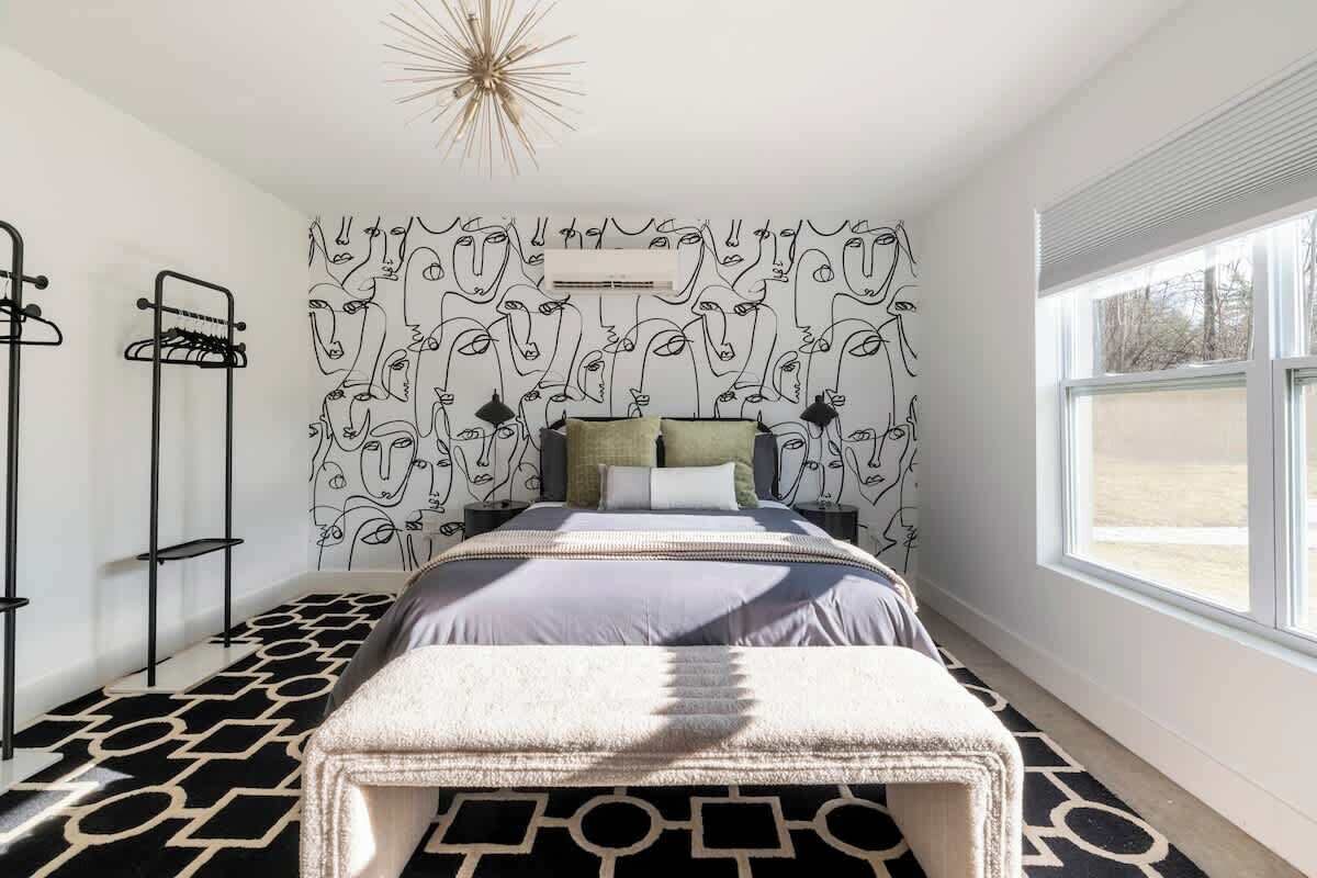 Une chambre avec un mur orné d’un motif, un lit surmonté d’un tapis à motifs géométriques, une fenêtre et un luminaire moderne.