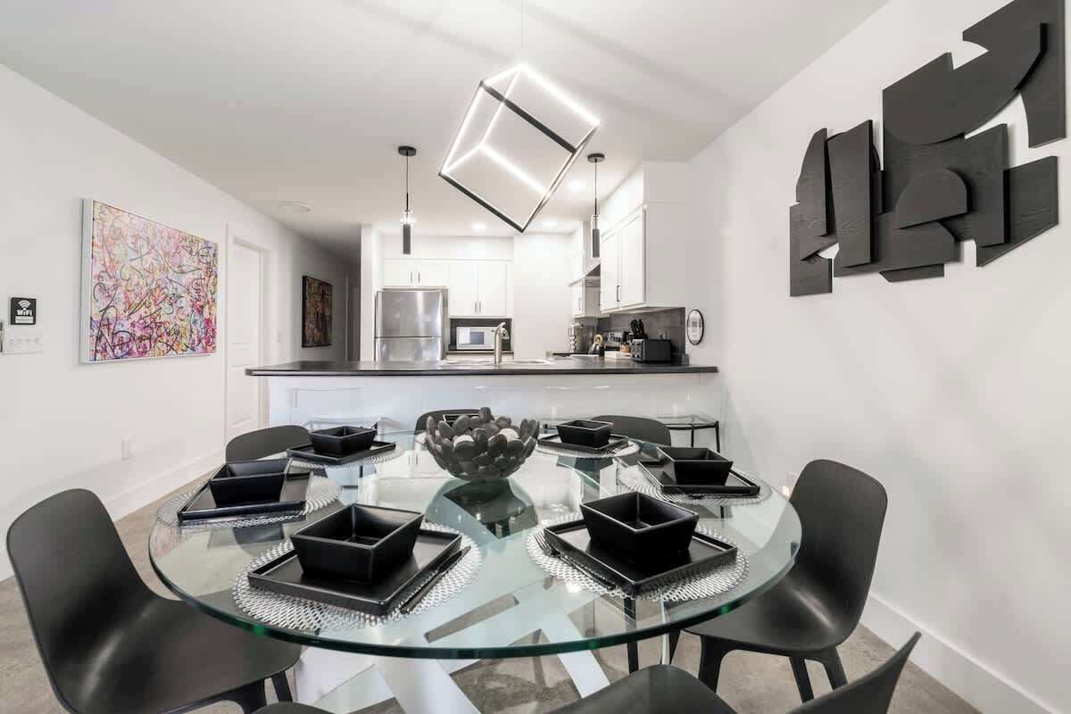Un espace repas moderne avec une table en verre entourée de chaises noires, un grand vase décoratif au centre et une cuisine en arrière-plan.