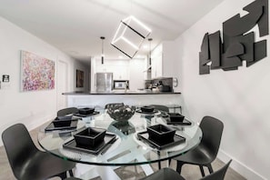 A modern dining area with a glass table surrounded by black chairs, a central decorative bowl, and a kitchen in the background.