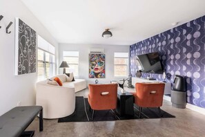 A modern living room with a white sofa, orange chairs, a black bench, and a TV on a wall with a circular pattern. - Gallery House: Designer Home-Hot Tub-Campfire-EV (Ithaca)