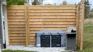 A wooden fence with a grill and a trash can in front. - Gallery House: Designer Home-Hot Tub-Campfire-EV (Ithaca)