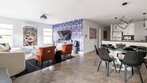 A modern living room with a sofa, orange chairs, a glass dining table, and a kitchen area with a refrigerator and cabinets. - Gallery House: Designer Home-Hot Tub-Campfire-EV (Ithaca)