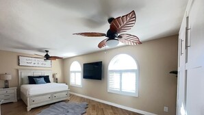 Un dormitorio con cama, ventilador de techo, televisor montado y un amplio ventanal.