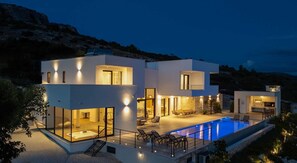 A modern two-story house with a swimming pool, outdoor seating, and a view of the surrounding landscape.
