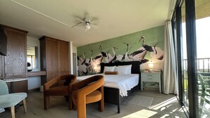 Une chambre à coucher comprenant un lit, deux chaises, un ventilateur de plafond, une table de chevet avec une lampe et une fresque murale représentant des oiseaux.