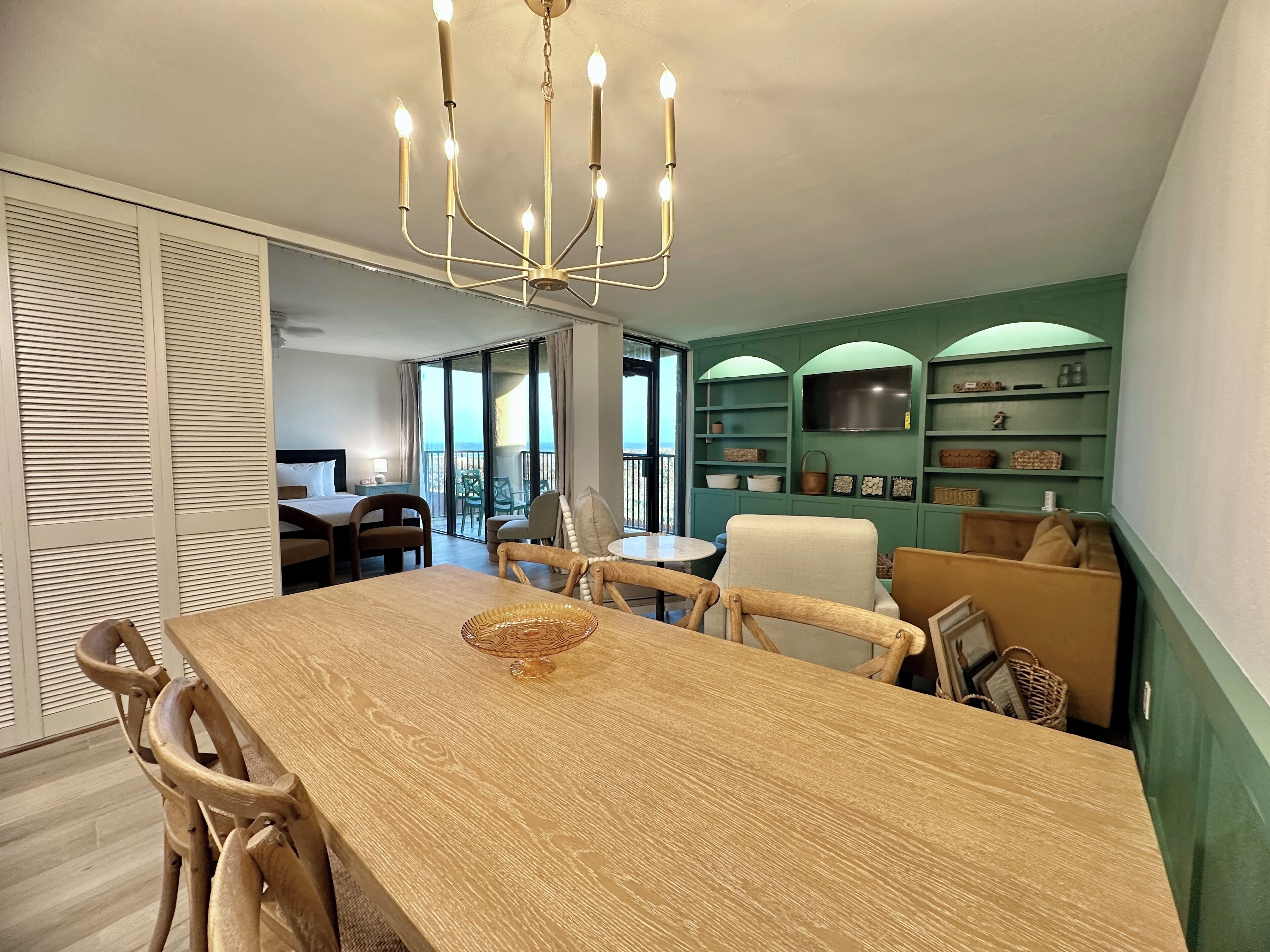 A dining area with a wooden table and chairs, a chandelier, and a view of the balcony.
