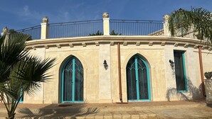 A building with a curved facade, two arched windows with teal frames, and a balcony with a metal railing. - Villa Brikinnia (Lentini)