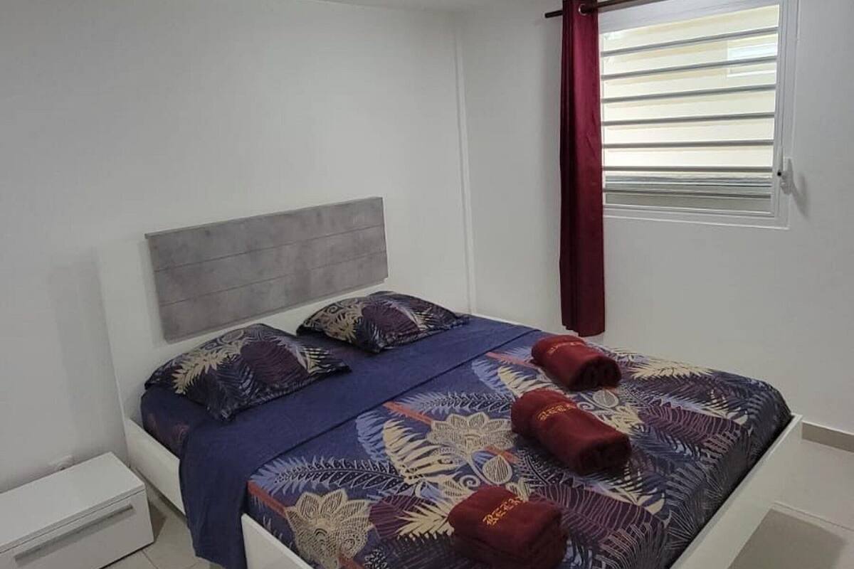 A modern bedroom with a bed, a small bedside table, a window with blinds, and a ceiling vent.