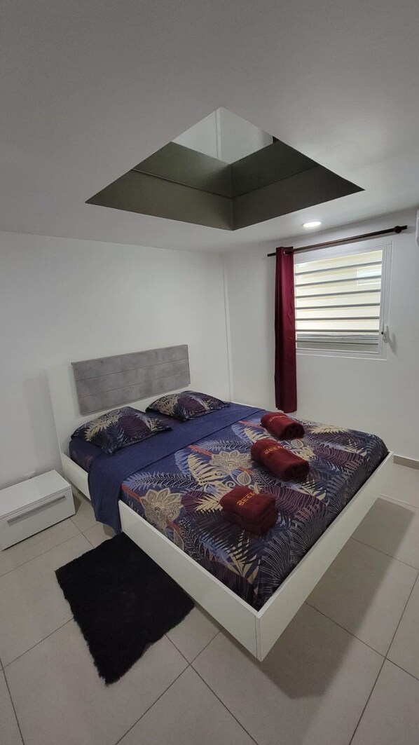 A modern bedroom with a bed, a small bedside table, a window with blinds, and a ceiling vent.