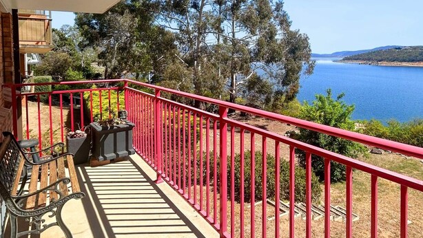 Un balcon avec une balustrade rouge donnant sur une étendue d’eau et un paysage vallonné.