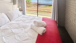 Un lit bien fait, avec du linge de lit blanc et un élément décoratif rouge, une fenêtre donnant sur un espace clos et un mur en briques.