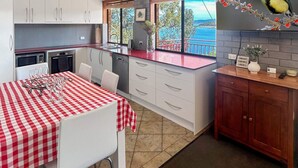 Une cuisine de taille modeste, avec une nappe à carreaux rouges et blancs, des placards blancs et une vue sur la mer par la fenêtre.
