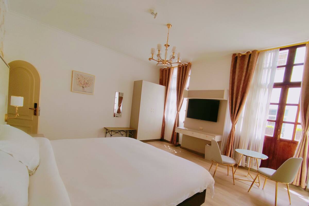 A hotel room with a large bed, a desk with a chair, a television, and a chandelier.