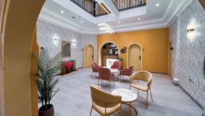Un vestíbulo amplio con techo alto, un área de descanso central con mesas redondas y una escalera que conduce a un nivel superior.