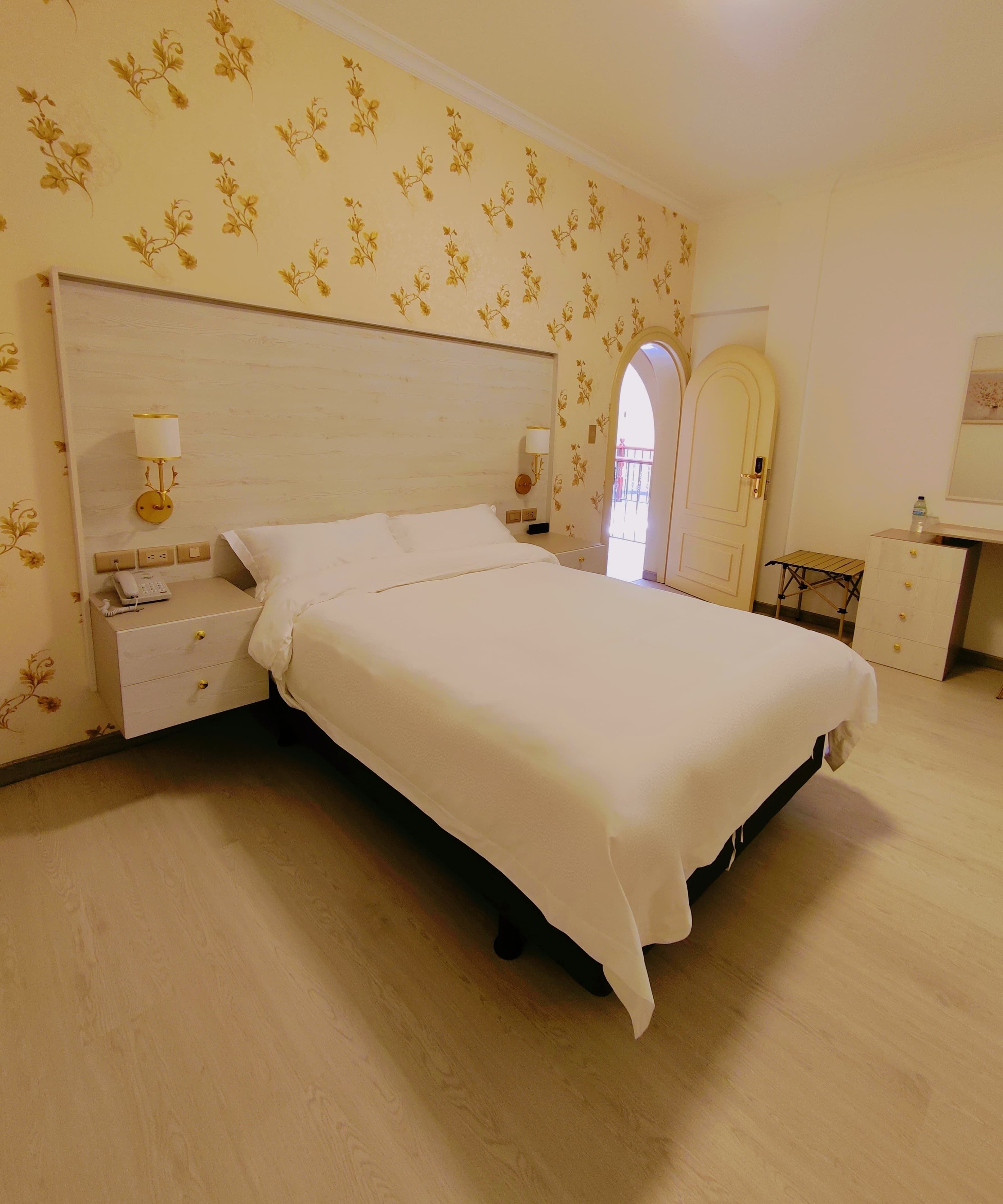 A neatly made bed in a hotel room with a floral wallpaper, a bedside table with a lamp, and a small desk with a water bottle.