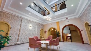 Un vestíbulo espacioso con techo alto, una zona de estar central con sillas rosas y una escalera que conduce a un nivel superior.