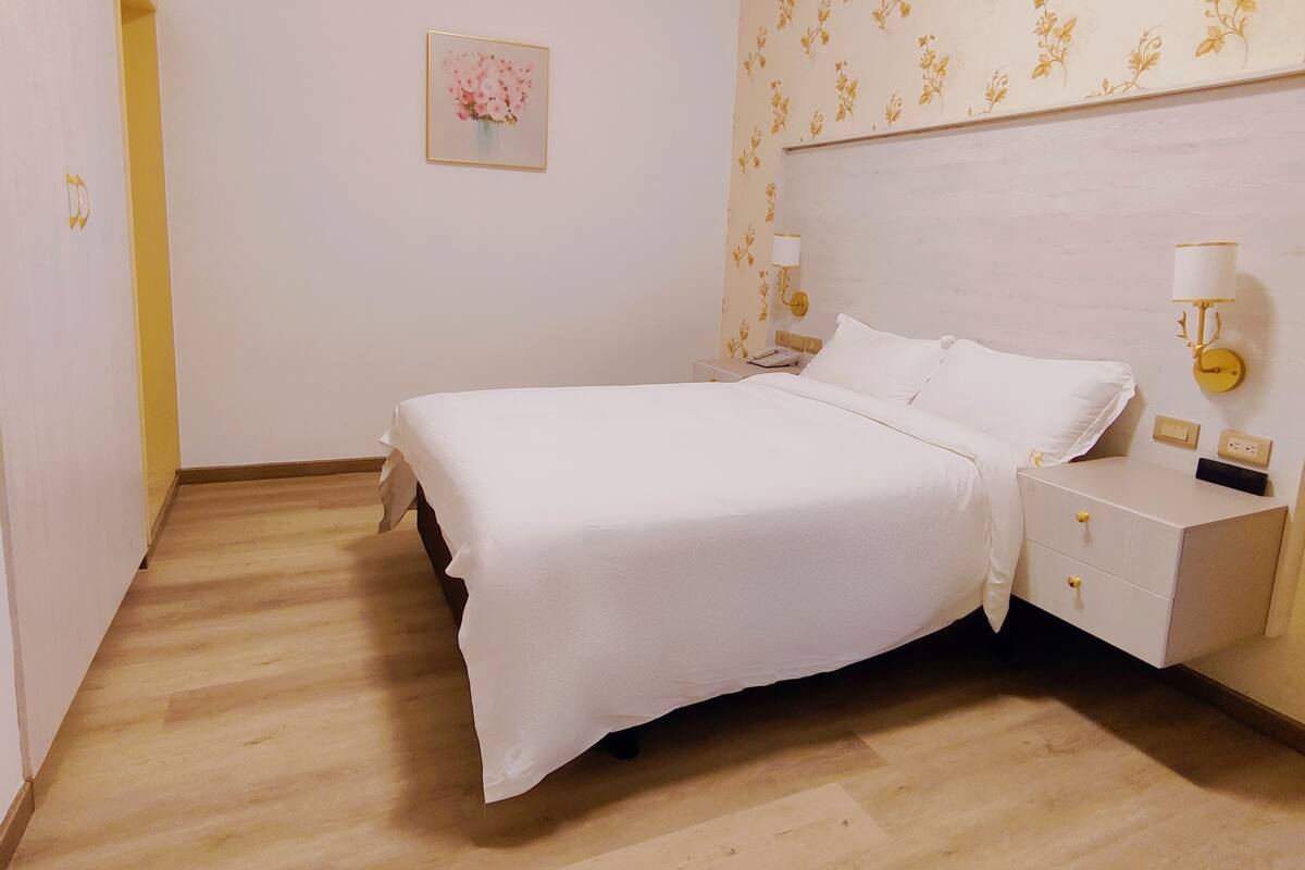 A bedroom with a bed, bedside tables, a nightstand, a wall-mounted lamp, and a floral wallpaper pattern.