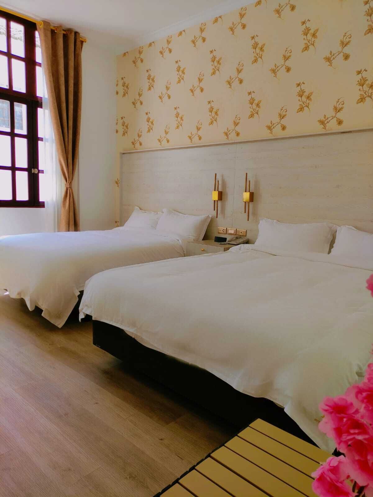 Habitación de hotel con dos camas, una ventana con cortinas, papel tapiz floral y piso de madera.