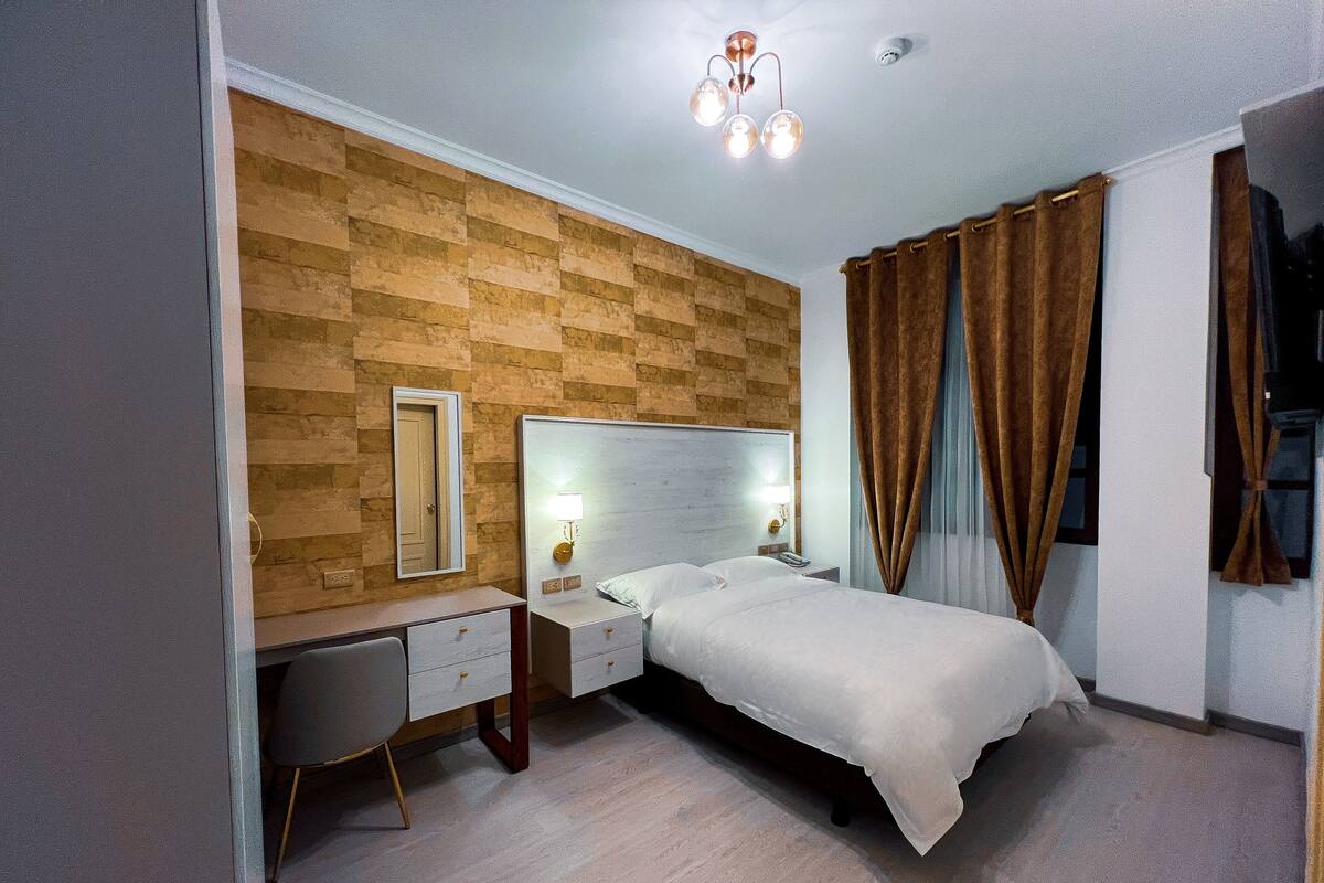 A hotel room with a wooden accent wall, a bed with white linens, a bedside table with a lamp, and a desk with drawers.