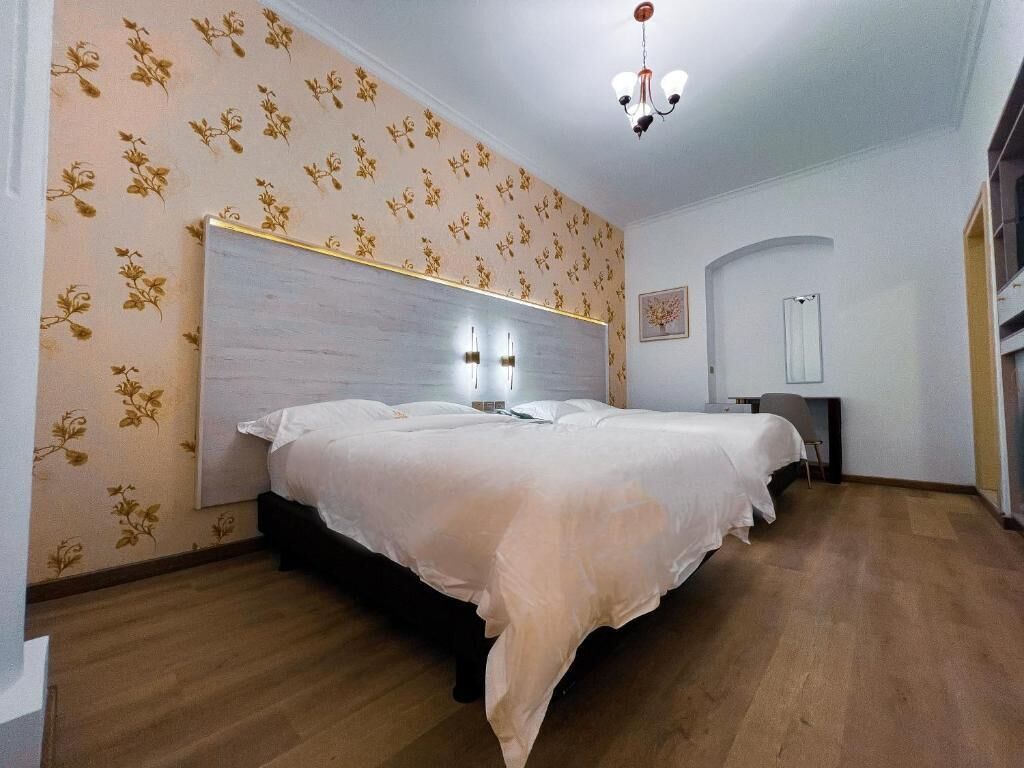 A bedroom with a large bed, wooden flooring, floral wallpaper, and a chandelier.