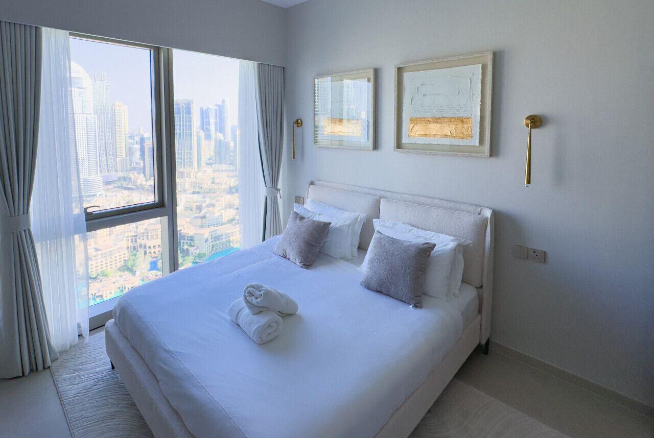 A hotel room with a large bed, two framed artworks on the wall, and a view of the city through the window.