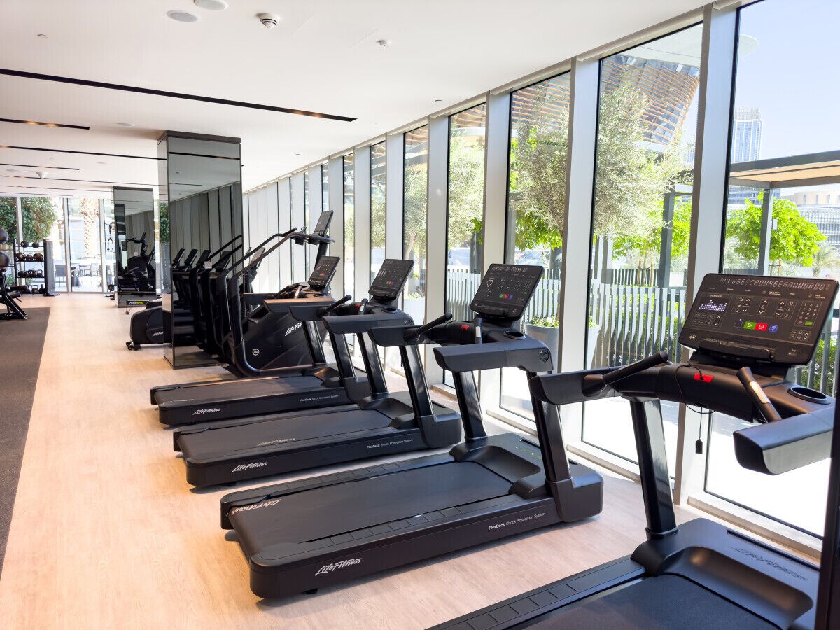 A row of treadmills in a modern gym with large windows and a view of the city.
