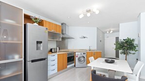A modern kitchen with a stainless steel refrigerator, wooden cabinets, a built-in oven, a washing machine, and a dining area with a table and chairs.