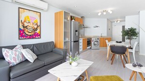 A modern apartment with a grey sofa, a dining table, a kitchen with a fridge, and a washing machine.