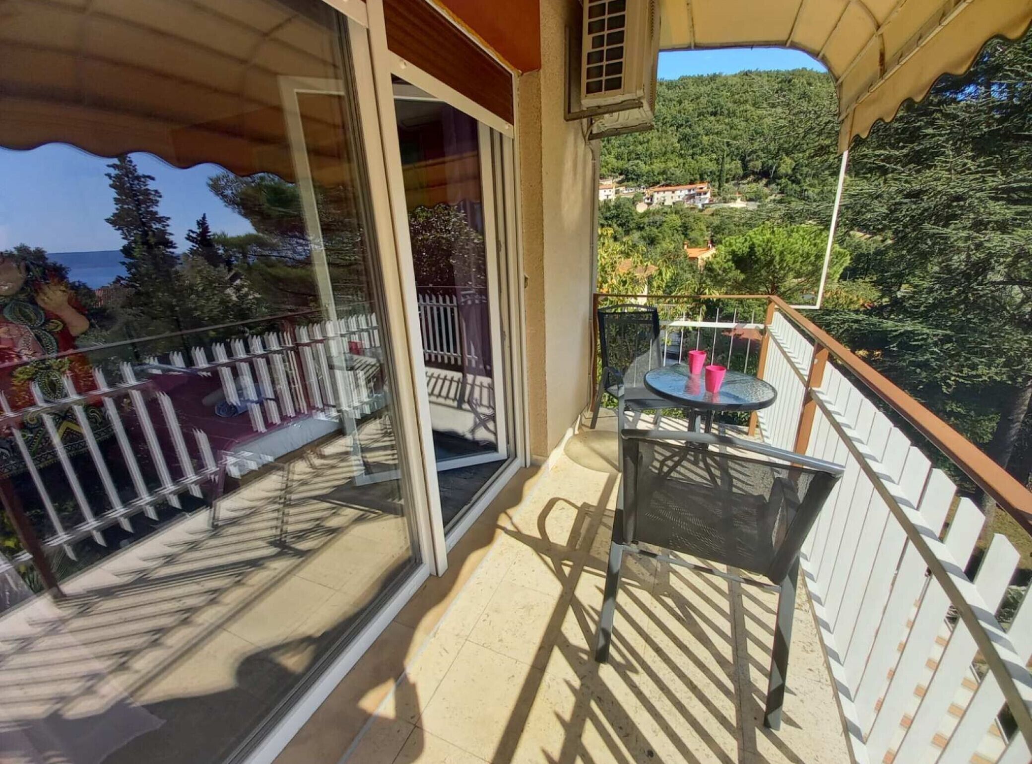 Ein Balkon mit Tisch und Stühlen, eine Glastür und Blick auf eine Hügelslandschaft mit Häusern.