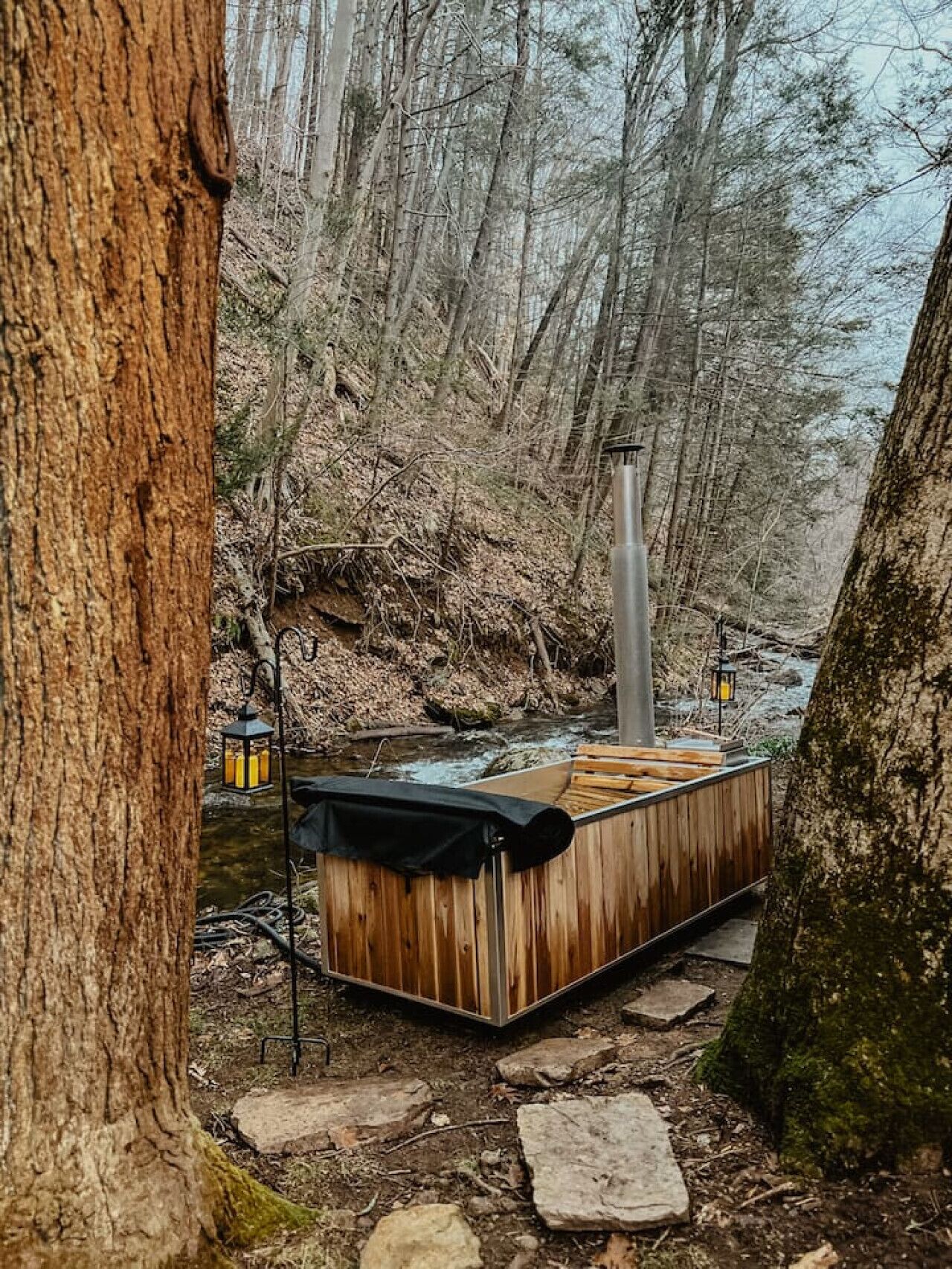 A wooden hot tub in a forested area with a stream, surrounded by trees and stone steps.