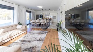 A spacious living area with a sofa, dining table, and chairs, a kitchen in the background, and large windows providing natural light.