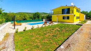 Une maison jaune à deux étages, dotée d’une piscine, d’une pelouse verdoyante et offrant une vue sur des collines en arrière-plan.