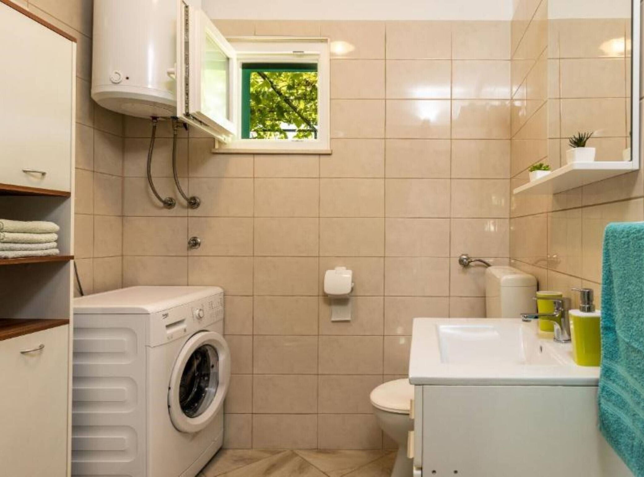 A compact bathroom with a washing machine, toilet, and a window with a view of greenery.