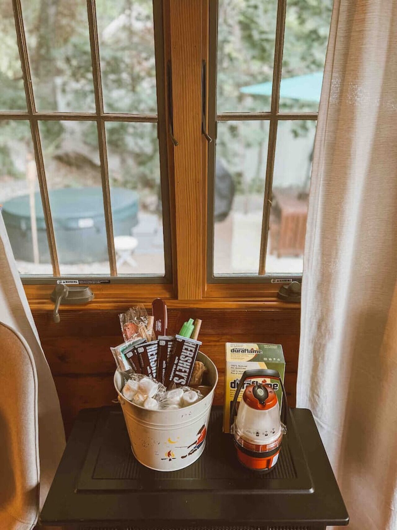 Une fenêtre donnant sur un paysage verdoyant, un seau rempli de barres de chocolat Hershey's et une bougie DuraFlame posée sur une petite table.