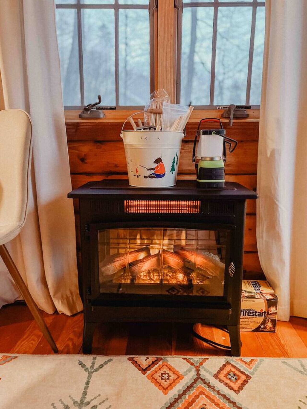 A cozy room with a wood-burning stove, a white bucket with a bear design, and a box labeled "Firestart".
