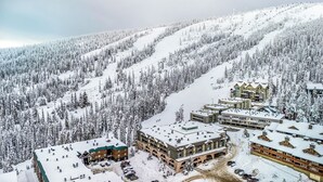 Ein schneebedecktes Bergresort mit mehreren Gebäuden, darunter eine Berghütte, im Hintergrund ist eine Seilbahn zu sehen.