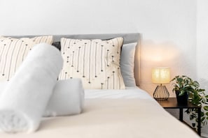 A neatly made bed with striped pillows and a rolled towel, a bedside lamp, and a potted plant on a small table beside the bed.