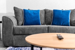 A grey striped sofa with two blue cushions, a wooden coffee table with a remote control, and a patterned rug.
