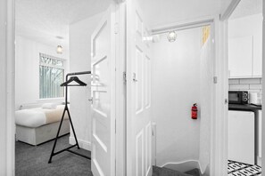 A modern, white-themed room with a bed, a hanging garment rack, a fire extinguisher, and a kitchenette.