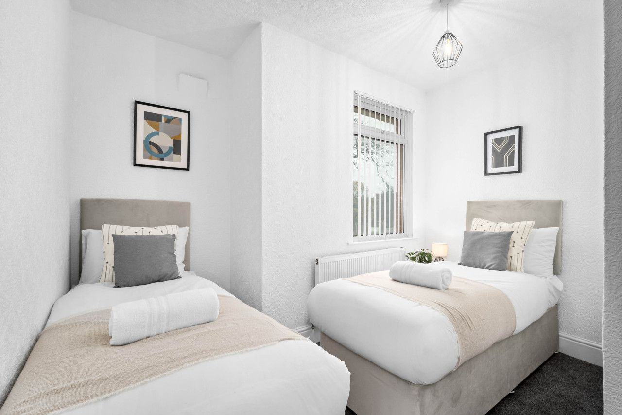 A modern, minimalist bedroom with two beds, white walls, and a large window with blinds.