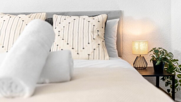 Ein ordentlich bezogenes Bett mit gestreiften Kissen und einem aufgerollten Handtuch, eine Nachttischlampe und ein Blumentopf auf einem kleinen Tisch neben dem Bett.