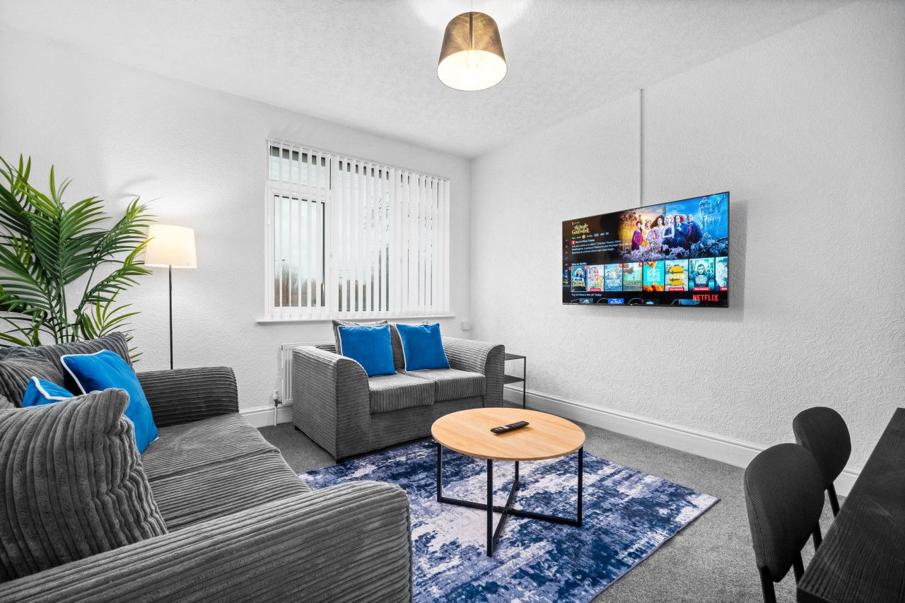 A modern living room with a grey sofa, blue cushions, a round coffee table, a TV mounted on the wall displaying a Netflix interface, and a large potted plant.