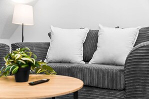 A grey upholstered sofa with white cushions, a small potted plant, and a remote control on a wooden coffee table.
