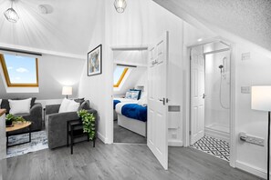 A modern, well-lit room with a grey sofa, a small table with a plant, and a bed with blue bedding.