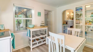 Een keuken met witte kasten, een houten aanrecht en een raam met jaloezieën. Er staat een witte eettafel met stoelen, en een wit kastje met glazen deuren waarin keukengerei staat.