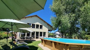 A wooden poolside area with lounge chairs and umbrellas, a two-story building with large windows, and a clear blue sky. - Gîte le Quai (8 people) (Champagnac-la-Rivière)